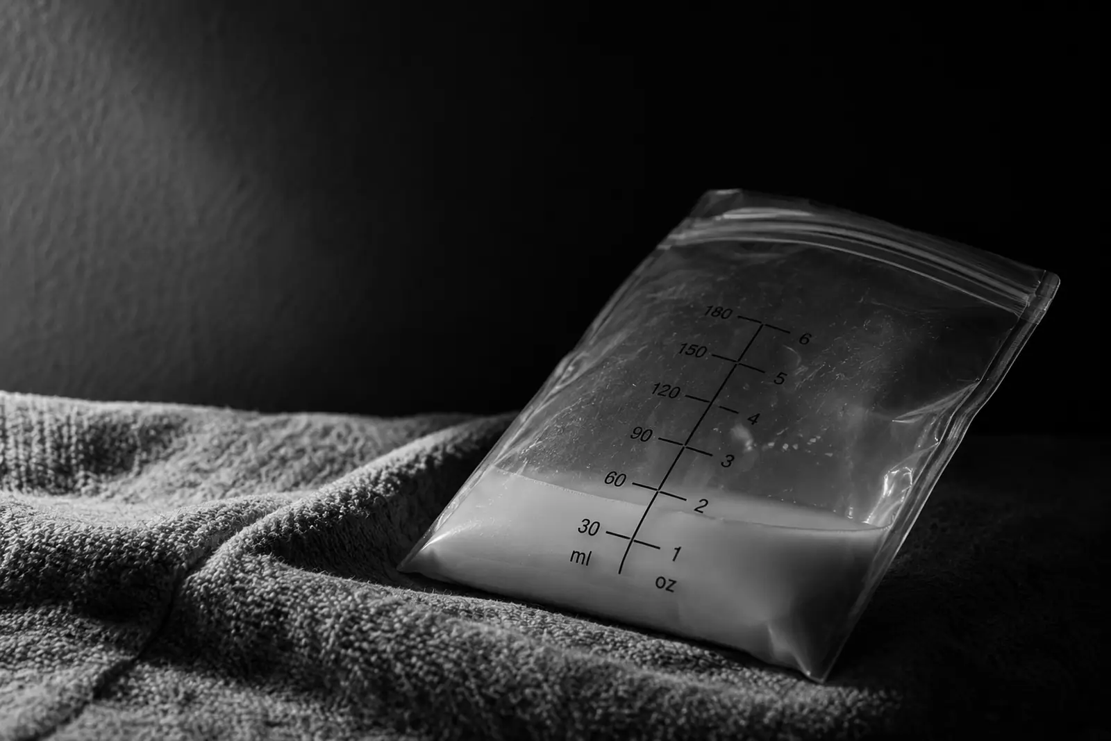 Gros plan en noir et blanc d'un sachet de conservation du lait maternel posé sur une couverture tricotée sur fond sombre, rempli juste en dessous de la ligne 60 ml / 2 oz de son échelle graduée imprimée.